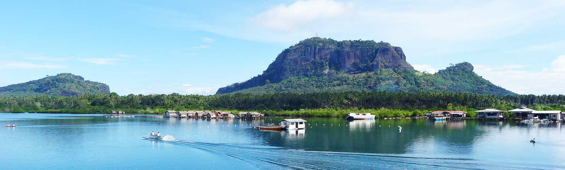 Scenic waterfront village with traditional stilt houses beneath volcanic mountain peak in tropical Philippines landscape